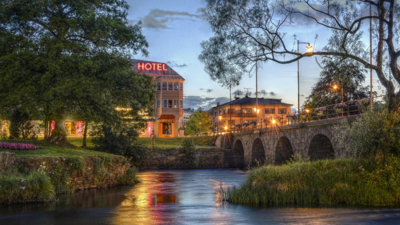 Charming twilight view of a hotel and stone bridge over a serene river with glowing lights and lush greenery.