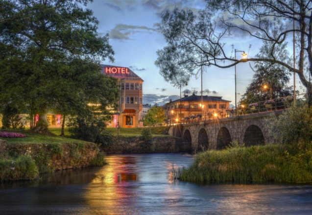 Charming twilight view of a hotel and stone bridge over a serene river with glowing lights and lush greenery.