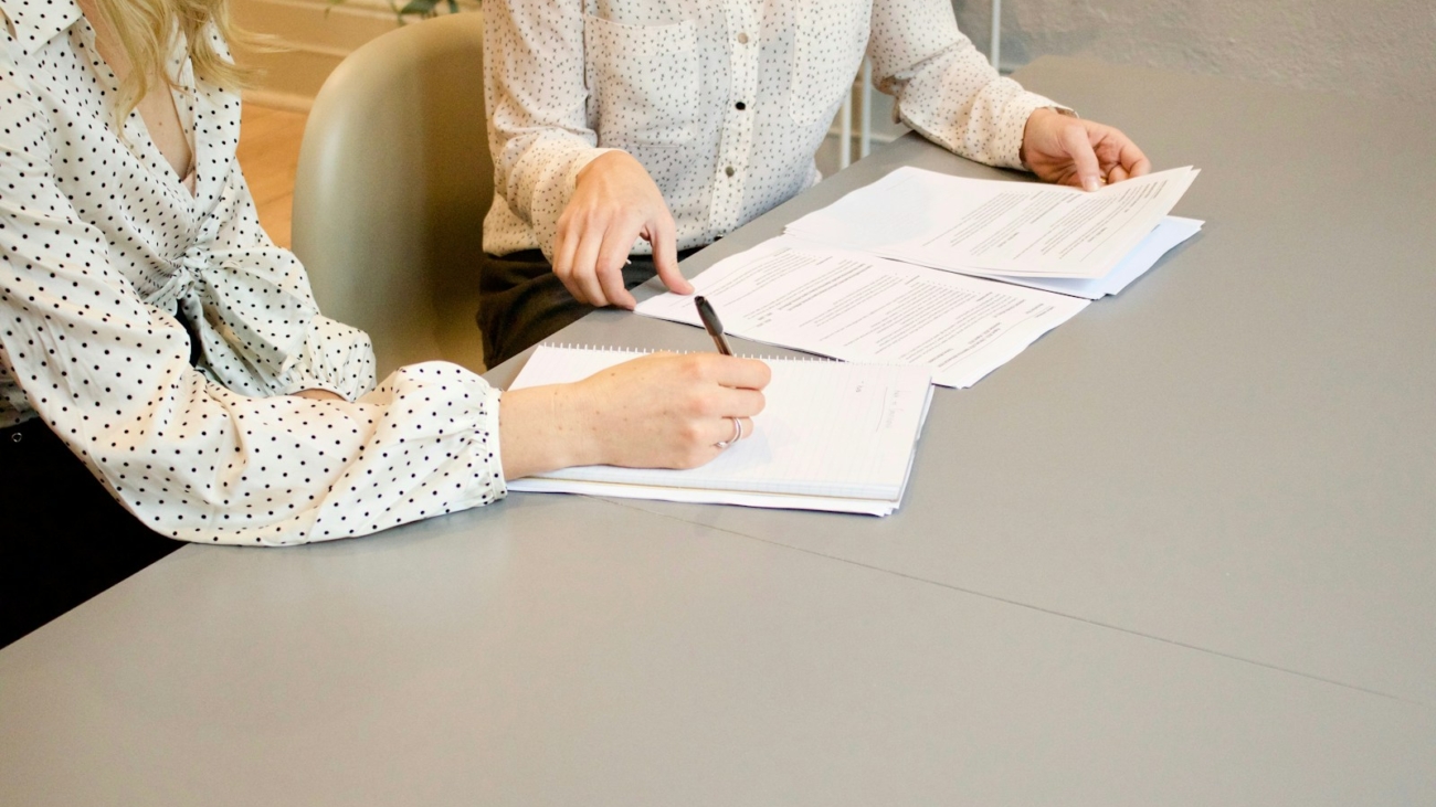 woman signing on white printer paper beside woman about to touch the documents